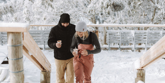 Woman and man walking in snow with coffee. Woman wears grey merino wool thermals and snow rust coloured ib pant. Man wears black snow jacket and tan pants.
