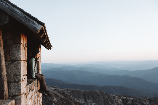 Lead image for blog "what does carbon neutral mean". Image is of girl sitting in window of stone mountain hut with landsape of blue toned mountains in the background.